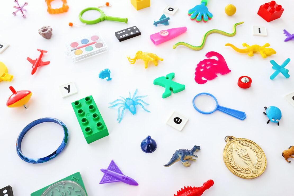 An assortment of colorful small toys and objects, including plastic animals, letters, a magnifying glass, a toy airplane, a spinning top, and building blocks, scattered on a white background