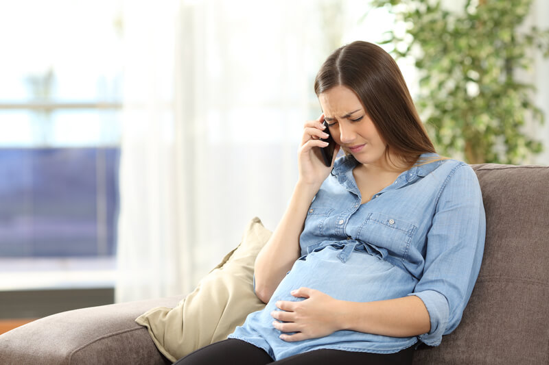 A person sitting on a couch, holding a phone to their ear with one hand and resting the other hand on their pregnant belly. They are wearing a light blue button-up shirt and black pants. A beige pillow is beside them, with green plants and bright natural light in the background