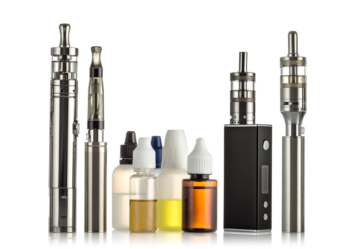a variety of four different silver vape pens and e-cigarettes with five clear bottles containing different colours of vape juice.