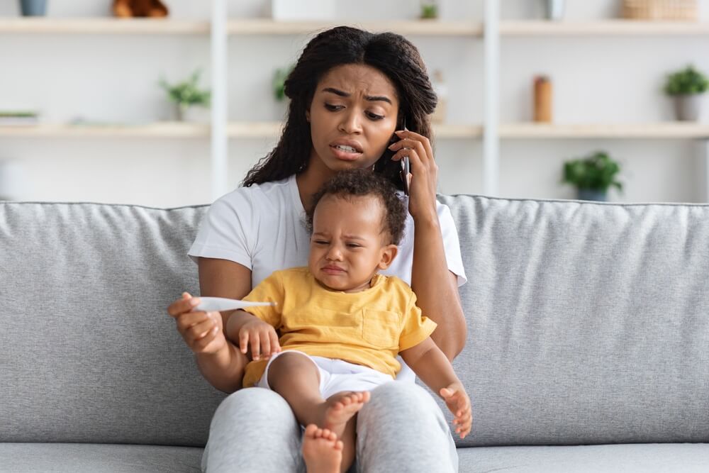An adult sitting on a gray couch, holding a child on their lap. The adult is wearing a white shirt and gray pants, and the child is dressed in a yellow shirt and white shorts. The background includes shelves with plants and decorative items